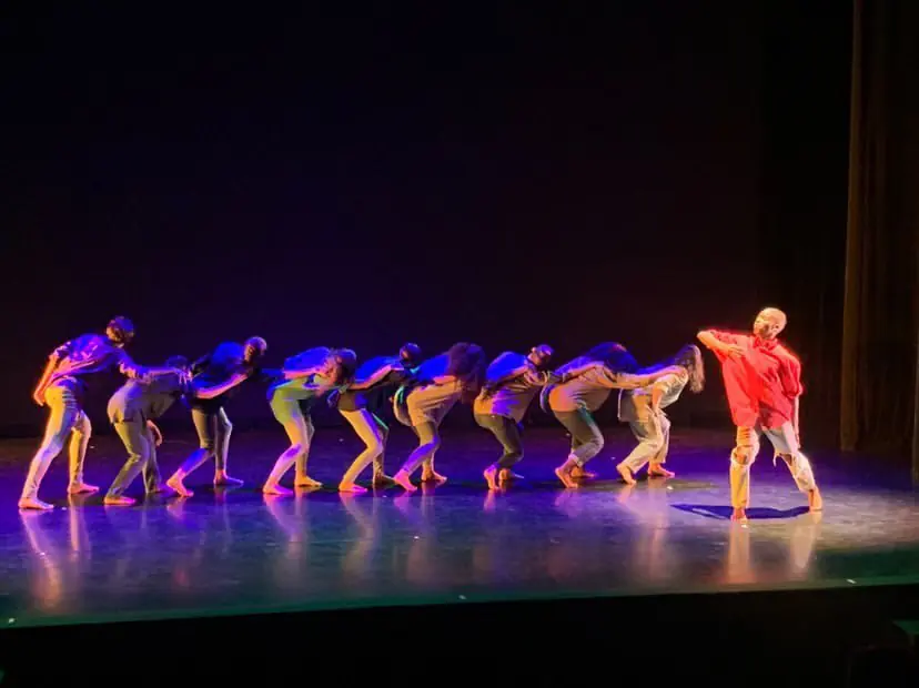 A line of dancers bowing forward on stage while a soloist in red stands apart