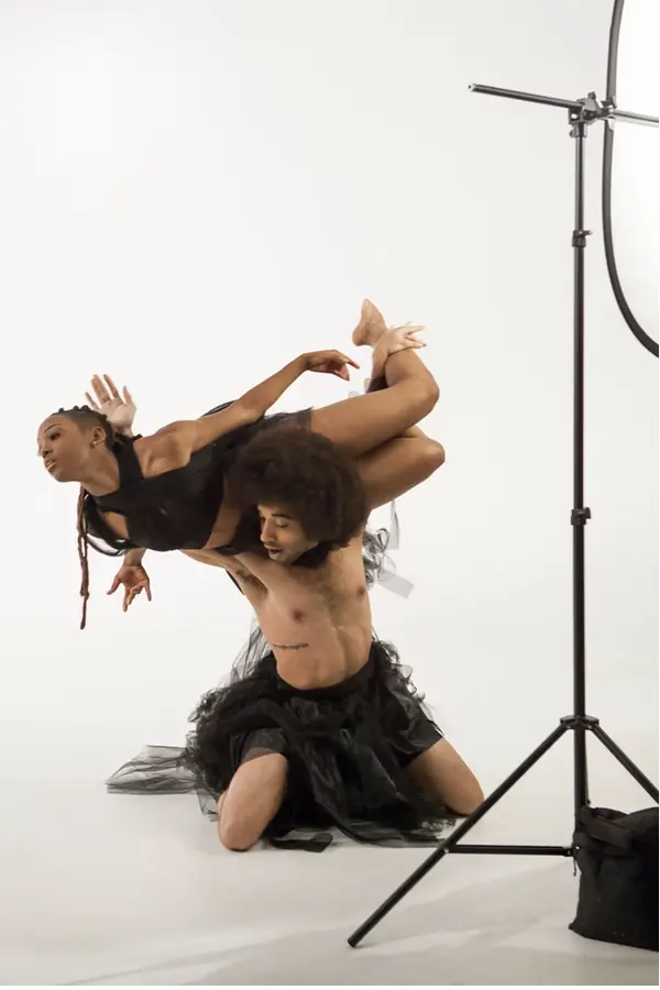 JustBee lifted across a partner's shoulders in black tulle