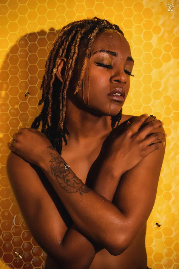 JustBee with honey on her face against a honeycomb wall, arms crossed