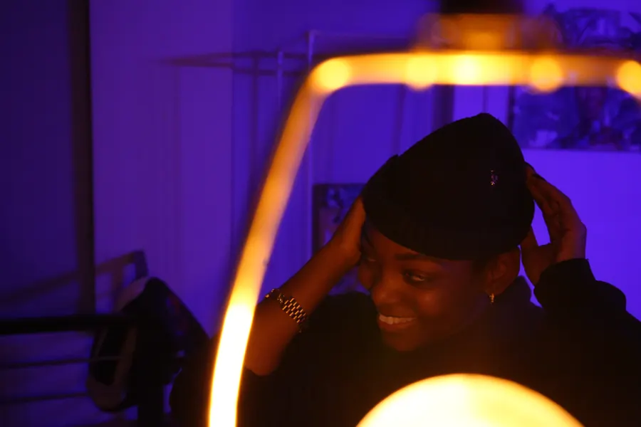 JustBee smiling in a purple-lit room, adjusting her beanie