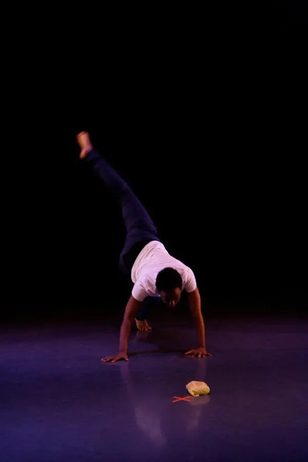 A solo dancer balanced on her hands, one leg raised, on a dark stage